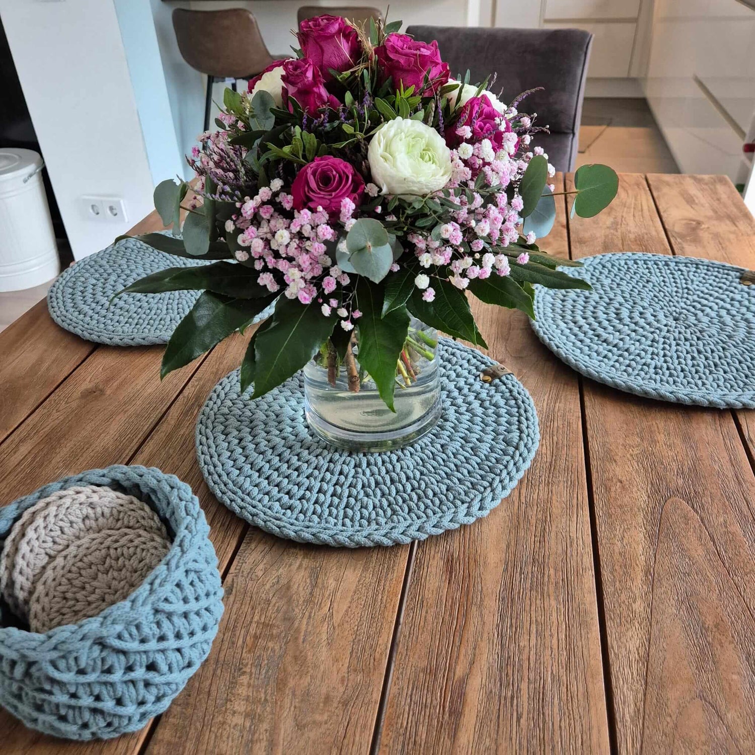 Gehäkelte Platzsets & Untersetzer aus recycelter Baumwolle in Blau auf Holztisch, dazu Blumenvase und kleiner Korb.